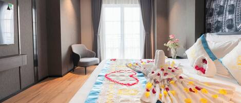 A hotel room with a bed adorned with a heart-shaped towel arrangement, a blue and white striped runner, and a vase with pink flowers.
