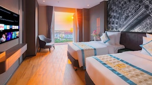 A hotel room with a TV displaying various channels, a balcony with a view of the sunset, two beds with blue and white bedding, and a large black and white artwork on the wall. - Times Corner Amalfi Hotel (Phu Quoc)