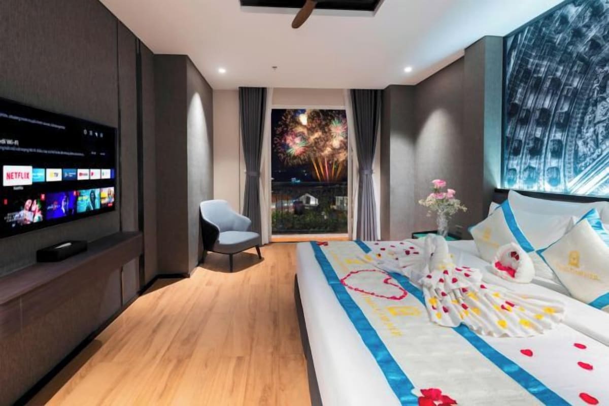 A modern hotel room with a large bed, a flat-screen TV mounted on the wall, a comfortable chair, and a balcony with a night sky view.