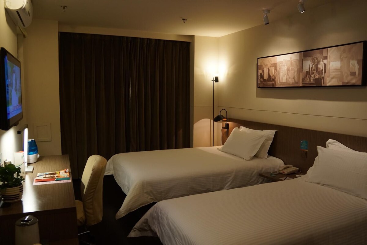 A hotel room with two beds, a desk with a computer and books, a TV, and a painting on the wall.