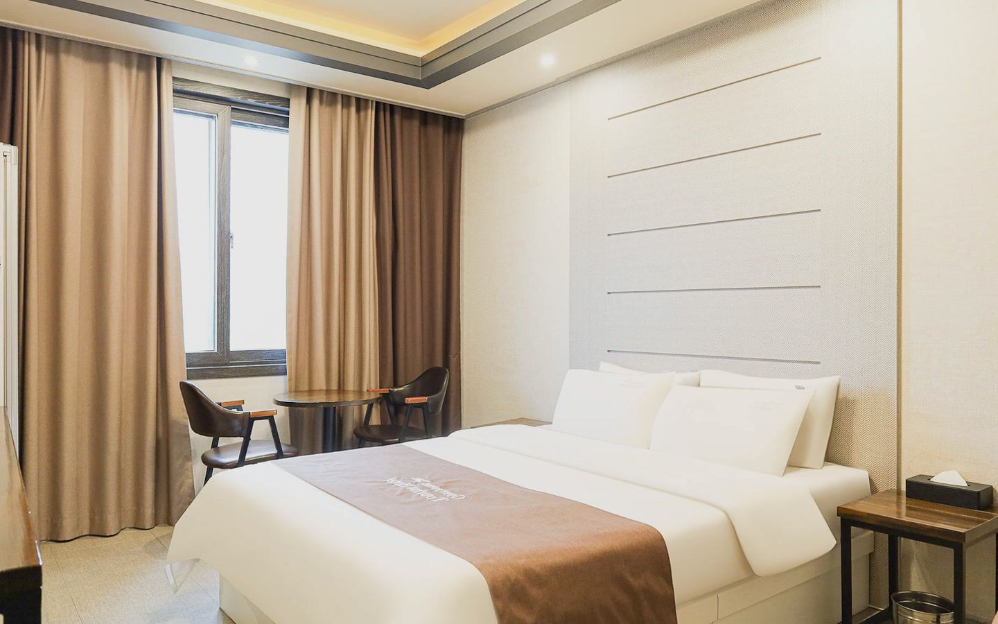 Una habitación de hotel con una cama, una mesita, dos sillas y un ventanal con cortinas.