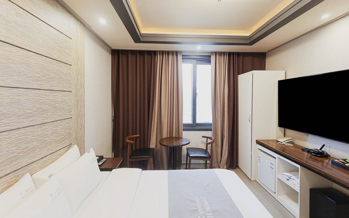 A hotel room with a bed, a desk, a television, a window with curtains, and a small table with chairs.