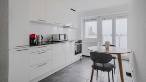 A modern kitchen with white cabinets, a black countertop, a microwave, a coffee maker, and a stove. There is a round table with two chairs and a view of buildings through large windows.