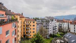 A row of multi-story residential buildings with balconies, a view of the cityscape in the background, and a cloudy sky.
