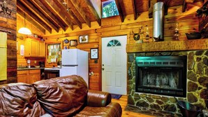 A cozy cabin interior with a stone fireplace, wooden walls, a leather couch, and a kitchen area.