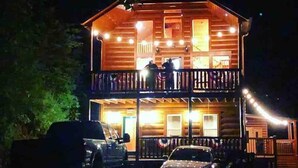 A two-story log cabin with a balcony, a porch, and string lights. - Beautiful Cabin w hot tub Sauna Pool Table (Sevierville)
