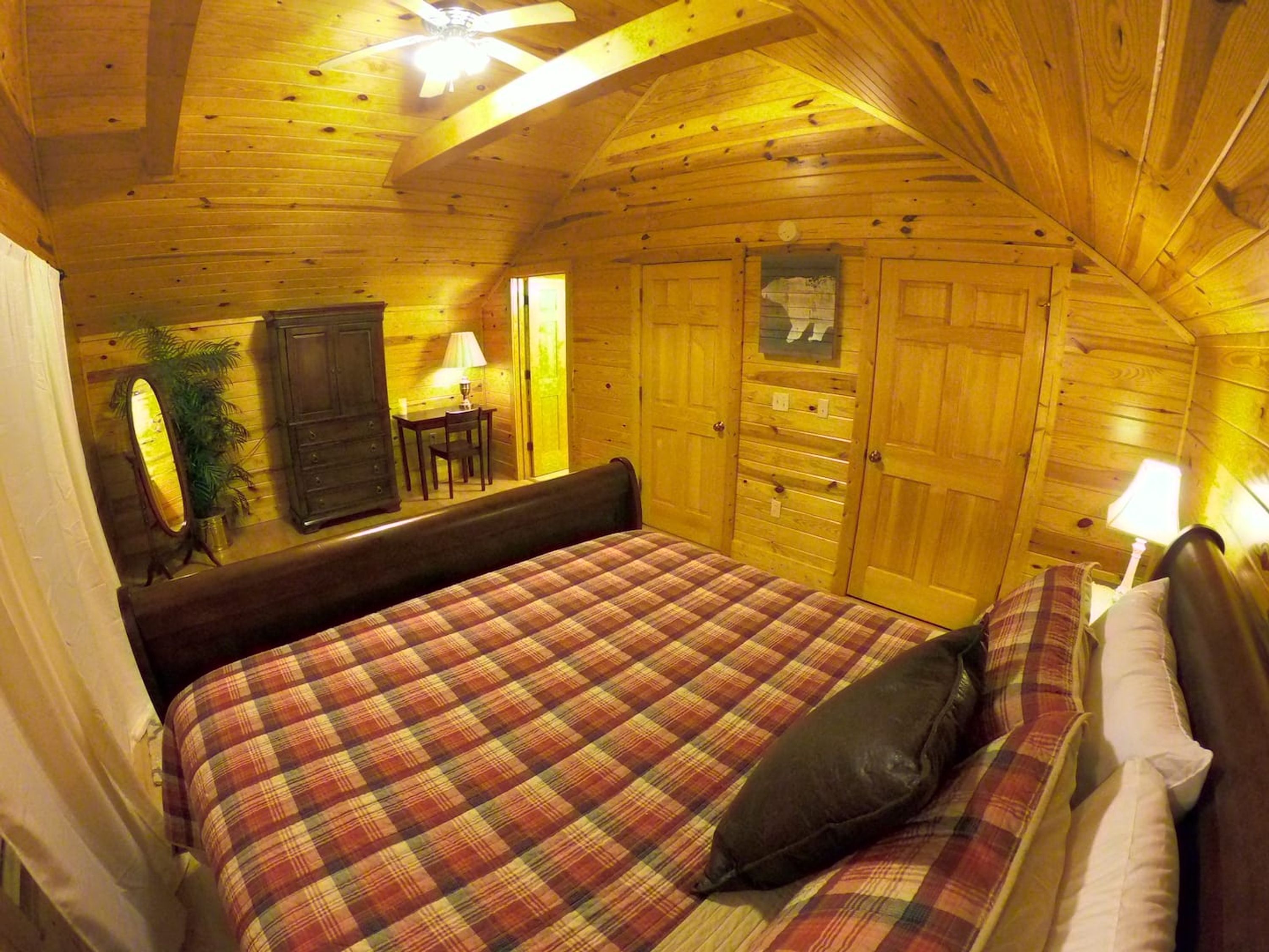 Een gezellig houten slaapkamer met een geruit sprei, houten wanden, een plafondventilator en een bureau met een lamp.