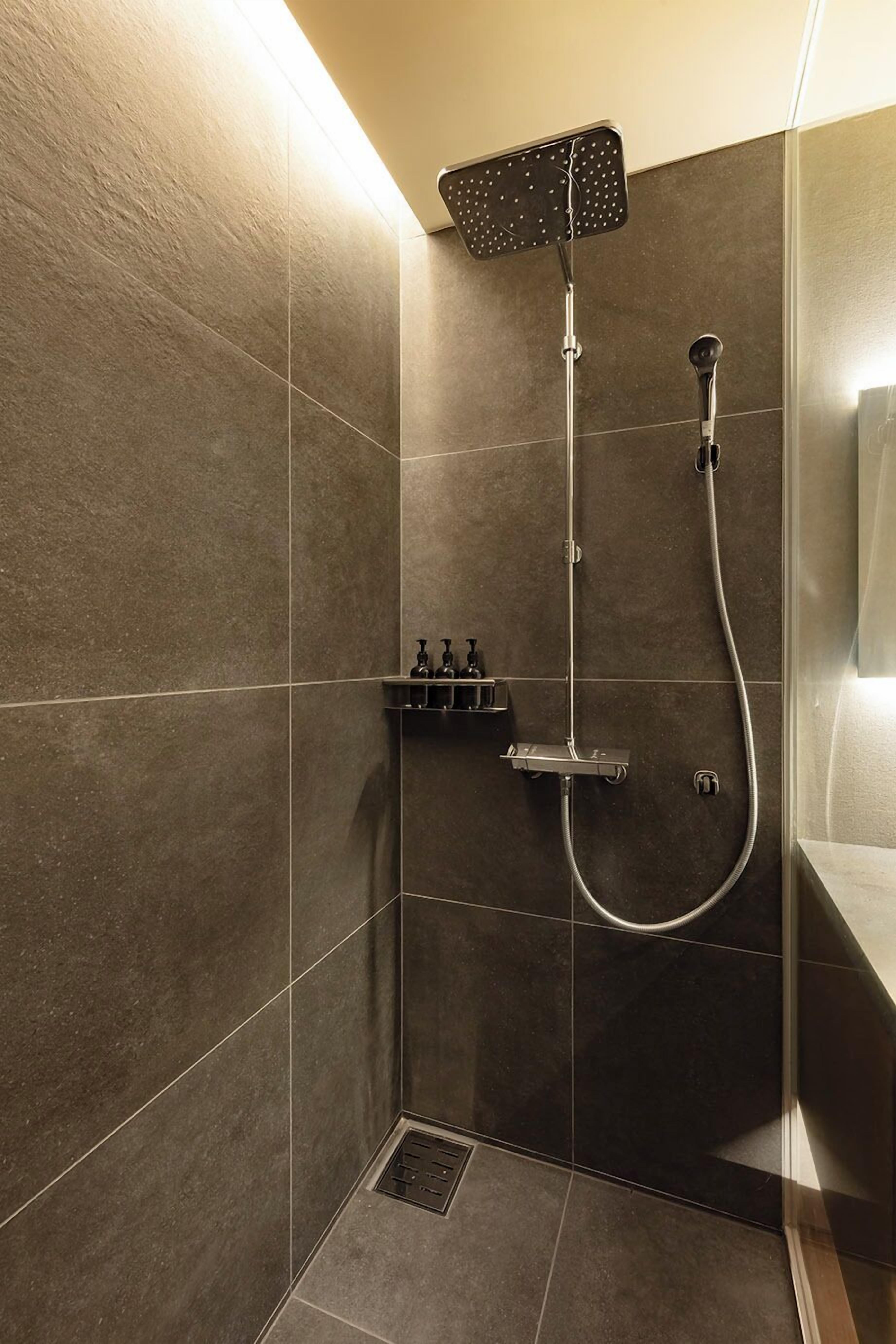 A modern shower with a handheld showerhead and a fixed showerhead, a control panel, and a small shelf with toiletries.