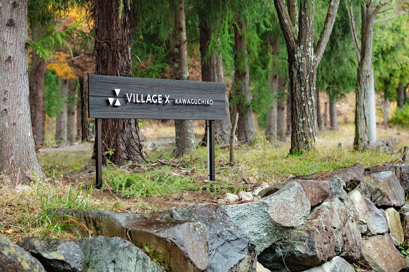 Un letrero de madera con el texto "VILLAGE X KAWACHIKO" en un entorno forestal.