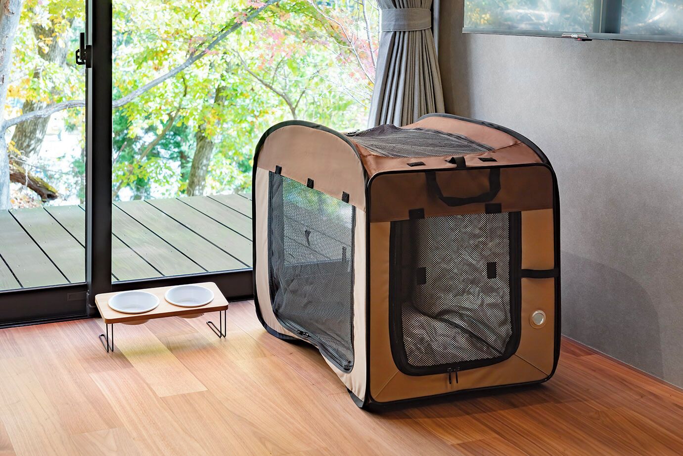A pet carrier in a room with a wooden floor, a small table with bowls, and a view of a wooden deck through a glass door.