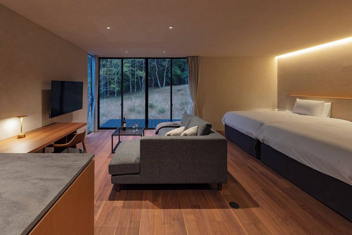 A modern hotel room with a wooden floor, a sofa, a bed, a desk with a lamp, and a TV.