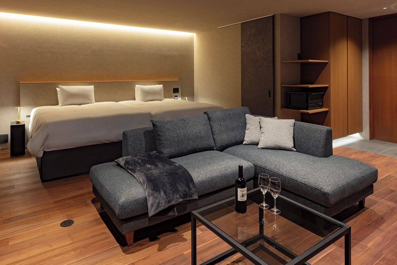 Ein modernes Hotelzimmer mit einem großen Bett, einem grauen Sofa, einem Glastisch mit Weingläsern und einer Flasche sowie einem Fernseher auf einer Holzablage.