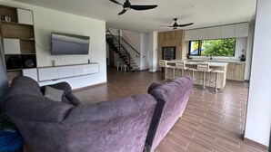 A modern living room with a large purple sofa, a flat-screen TV mounted on the wall, and a kitchen area with a bar and stools.