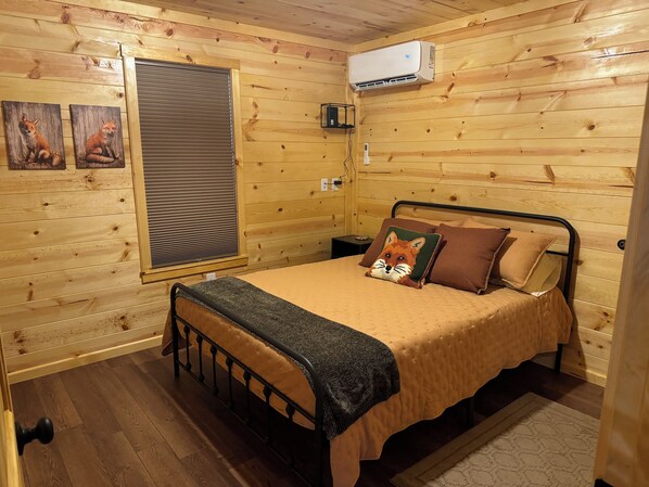 Un dormitorio acogedor de cabaña de madera con una cama, un ventanal con persianas, un equipo de aire acondicionado y dos cuadros con motivos de zorro en la pared.