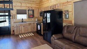 Interior de una cabaña de madera con cocina, un refrigerador negro, un sofá de cuero marrón y una araña.
