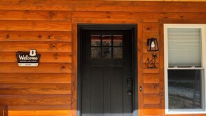 Una cabaña de madera con puerta negra, un cartel de bienvenida y una ventana con lámpara.