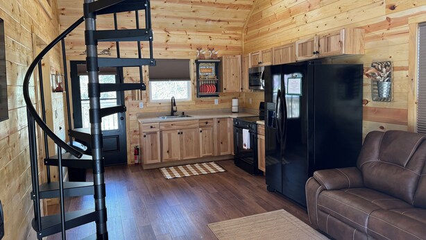 Un interior de cabaña acogedor con una escalera espiral, paredes de madera, una cocina con refrigerador negro y un sofá de cuero marrón.