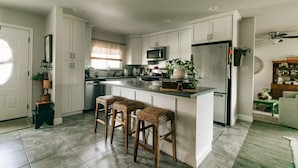 A modern kitchen with white cabinets, a central island with bar stools, a refrigerator, and a microwave. - Charming 2-bedroom house in quiet country Visalia with pool (Visalia)