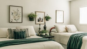 A bedroom with two beds, a bedside table with a lamp, framed pictures, and potted plants. - Charming 2-bedroom house in quiet country Visalia with pool (Visalia)