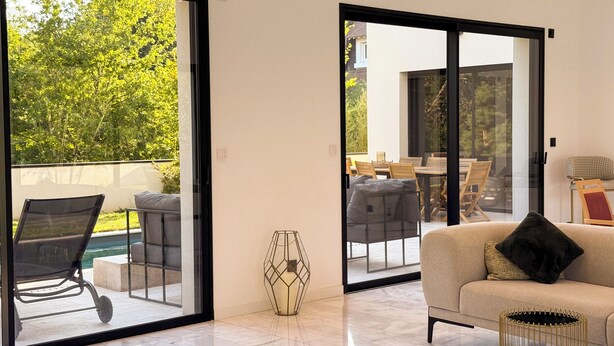Un saló modern amb un sofà, una taula de centre i una porta de vidre que dona a un pati exterior amb piscina i zona de menjador.