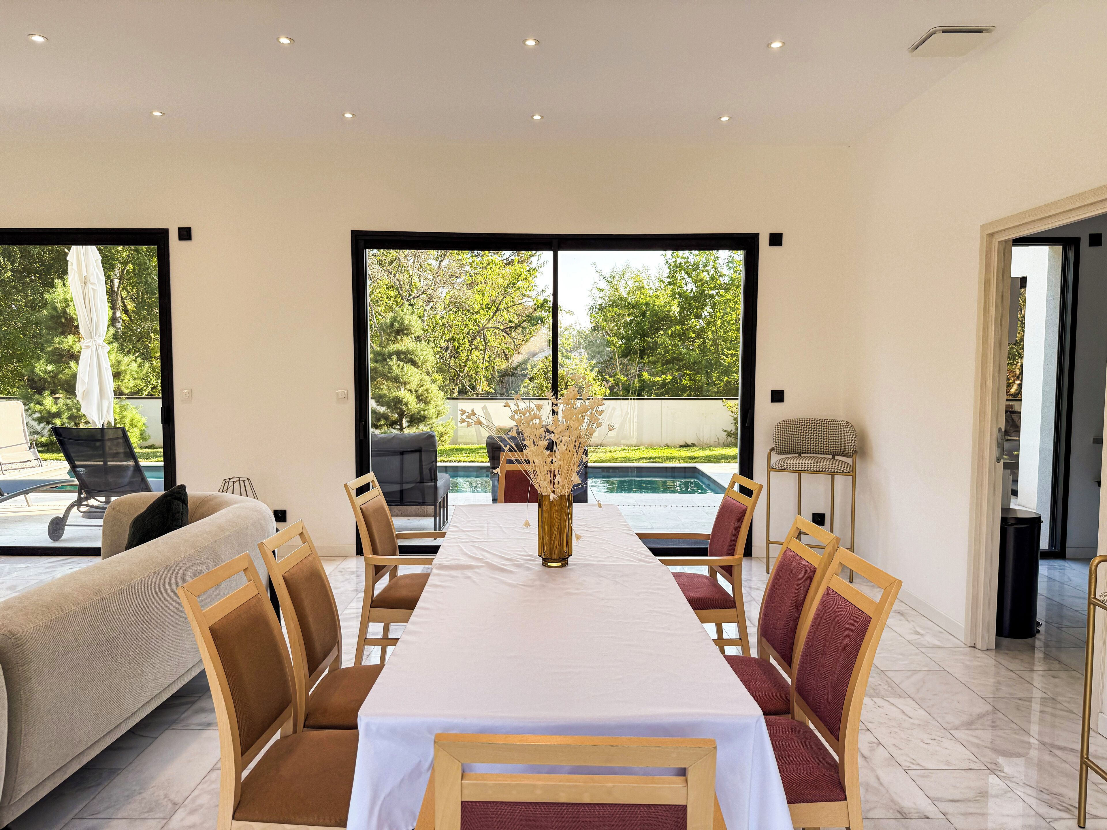 A dining area with a long table, chairs, and a view of a swimming pool and greenery outside.