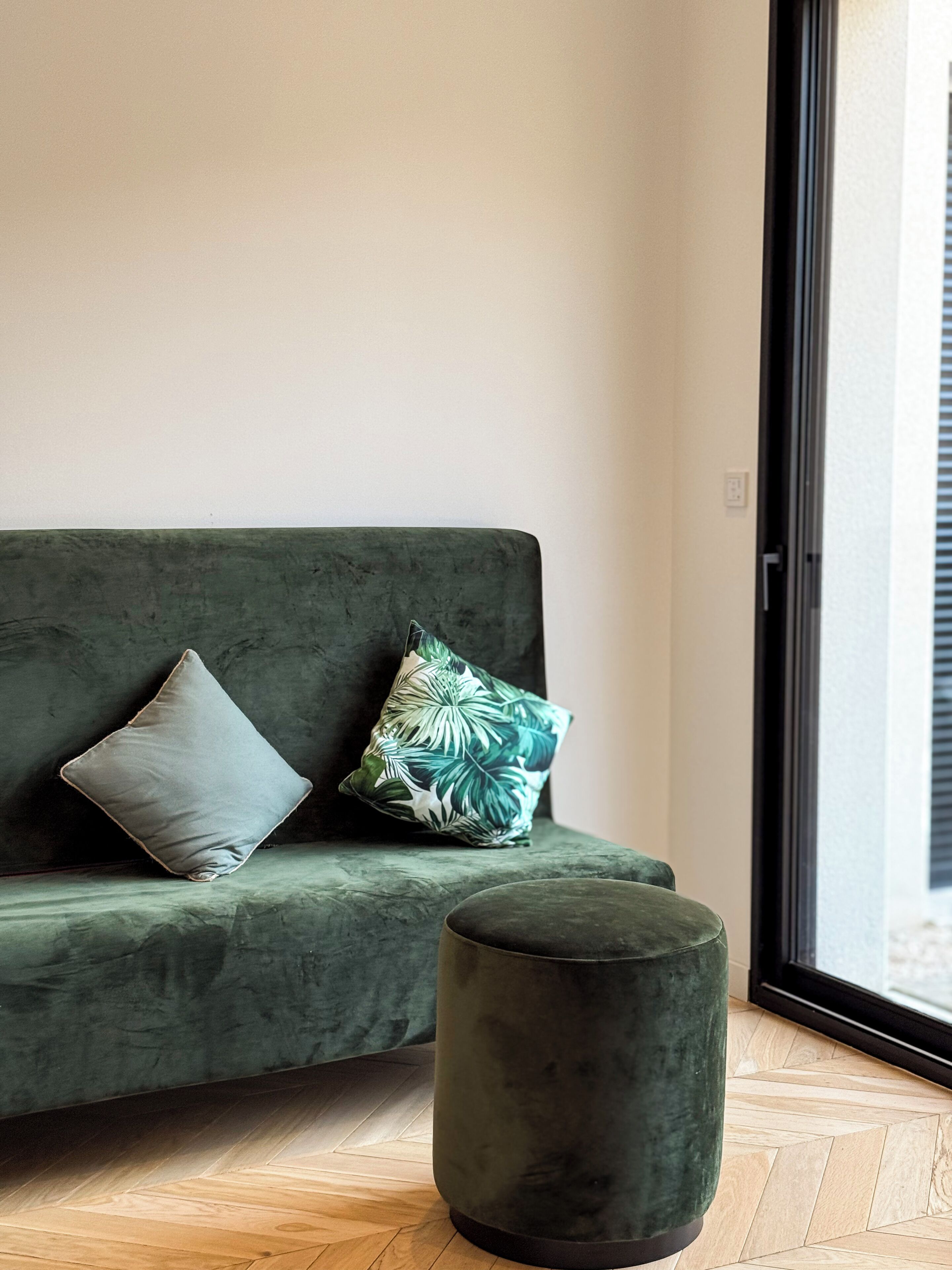 A green sofa with two pillows, one plain and one with a leaf pattern, and a matching ottoman in front of a window with blinds.