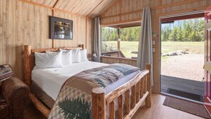 A wooden cabin bedroom with a bed, a wooden bench, and a large window offering a view of a forested area.