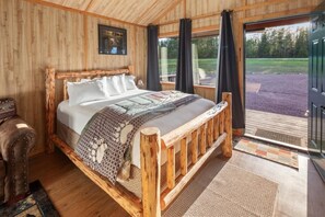 A wooden-framed bed with a patterned blanket, a wooden chair, and a view of a grassy area through a glass door. - Moose Cabin-6 Miles to Glacier! (Coram)