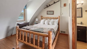 Un dormitorio con techo inclinado, estructura de cama de madera, mesita de noche, lámpara y vistas al exterior a través de una ventana.