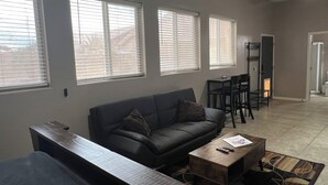 A living room with a sofa, coffee table, and a TV stand. There are three windows with blinds, a ceiling fan, and a door leading to a bathroom. - Cozy Guest House Apartment w/ Shared Backyard Oasis, Spa & Pool (Litchfield Park)
