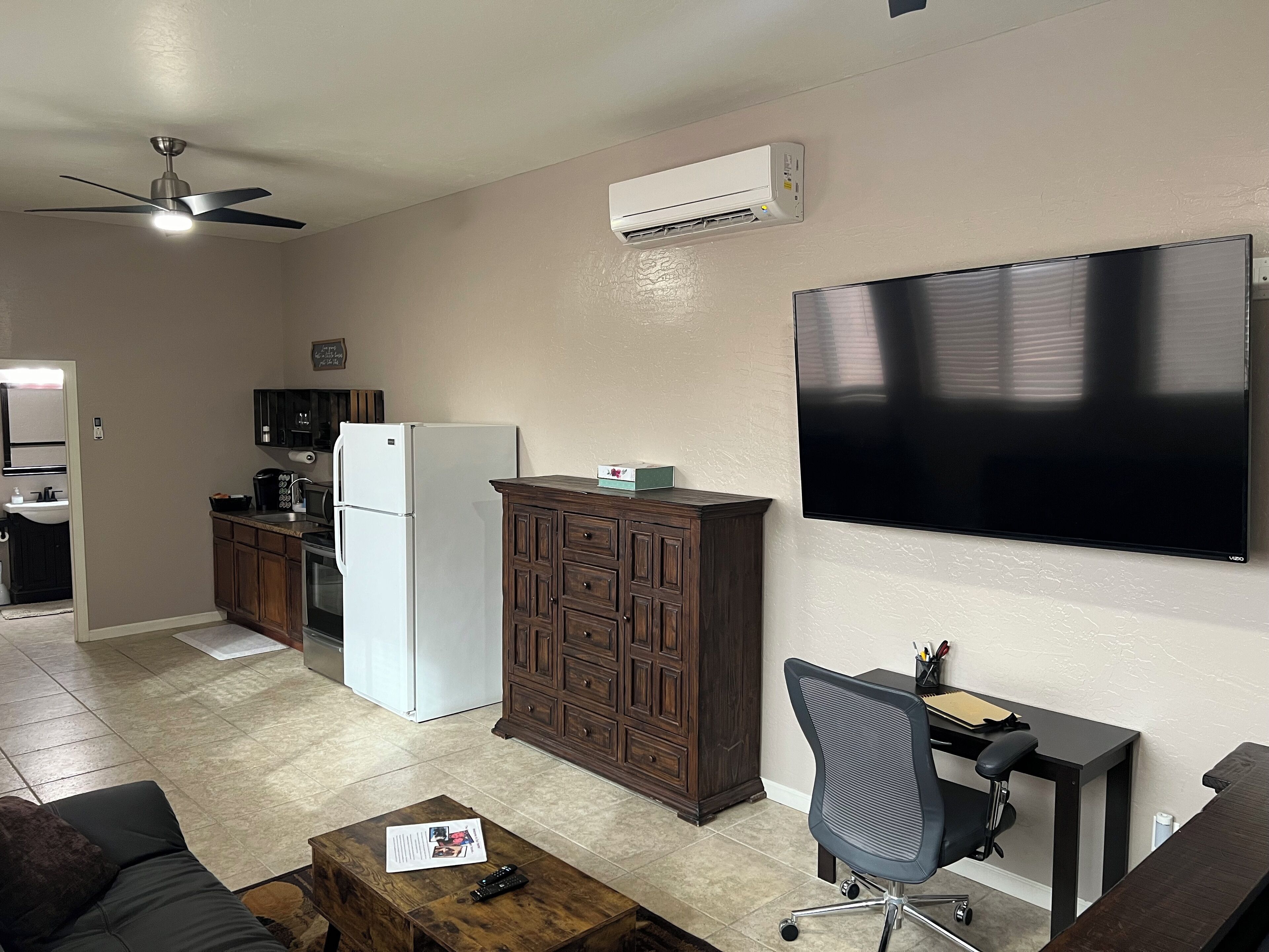 Een moderne woonkamer met een flatscreen televisie, een houten kast, een keuken met een koelkast en een plafondventilator.