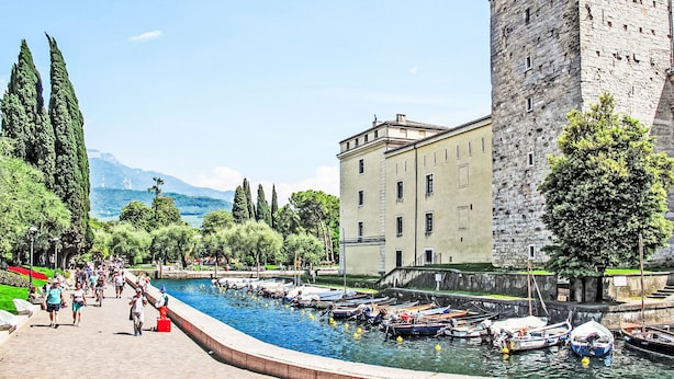 A historic stone building with a tower, a canal with boats, and a promenade with people walking.
