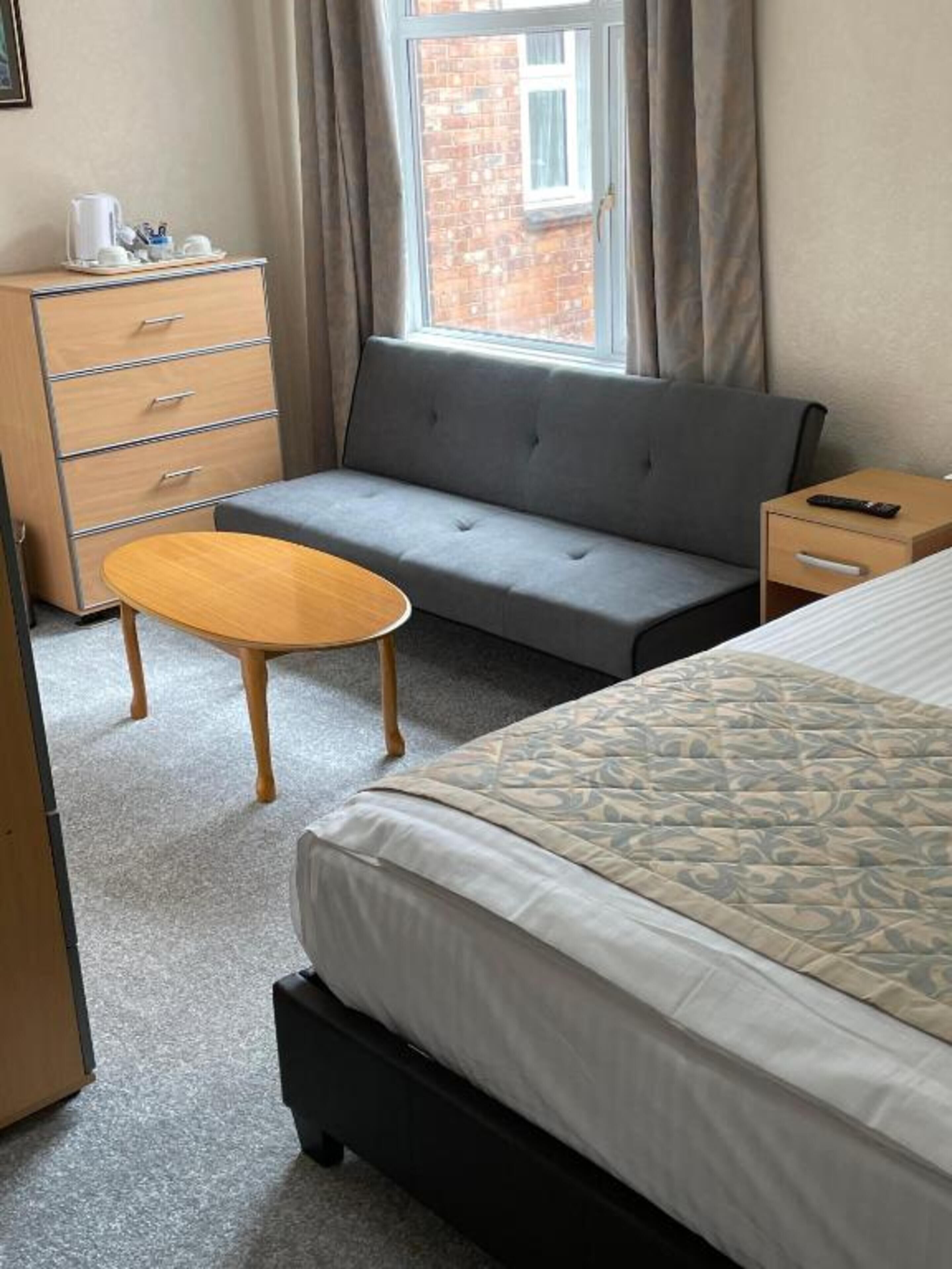 Una camera d'hotel con un letto, un divano grigio, un tavolo in legno, una cassettiera e una finestra con tende.