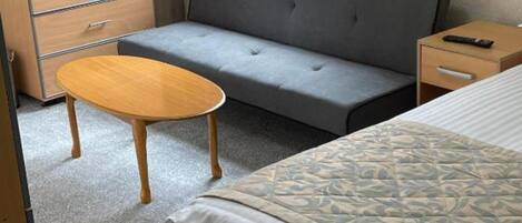 A hotel room with a bed, a grey sofa, a wooden table, a dresser, and a window with curtains.