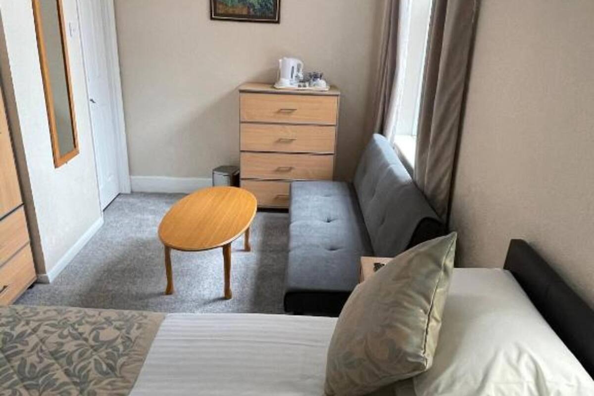A compact hotel room with a bed, a small wooden table, a grey sofa, a dresser, and a painting on the wall.