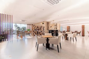 A modern hotel lobby with a round table and chairs, a wooden bookshelf, and a large window with a view of palm trees. - Hermosas vistas en Piso 10 en Camino al Mar (Mazatlán)
