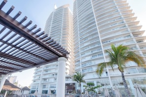 A high-rise hotel with multiple balconies, a pool area with umbrellas, and a palm tree. - Hermosas vistas en Piso 10 en Camino al Mar (Mazatlán)