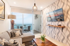 Un salon moderne avec un canapé, une table basse, un téléviseur fixé au mur et un balcon offrant une vue sur la mer.