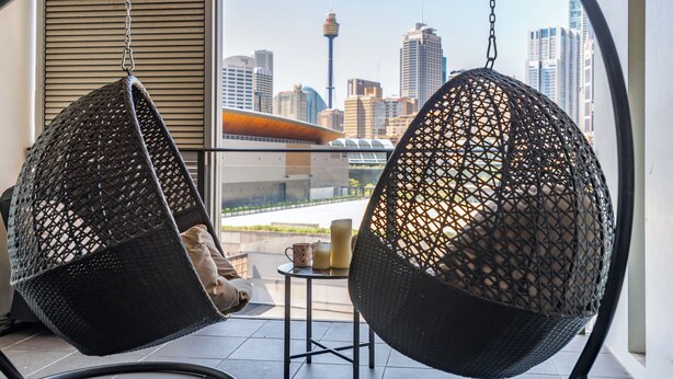 Dos sillones colgantes de mimbre negro con cojines, una mesita con velas y vista a un horizonte urbano con edificios altos y una torre.