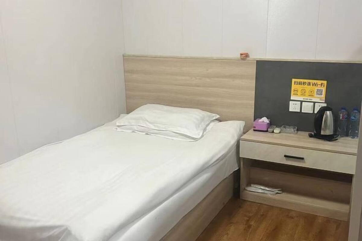 A single bed with a white mattress and pillow, a wooden headboard, a bedside table with a kettle, a small drawer, and a wall-mounted sign.
