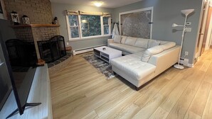 Een woonkamer met een stenen open haard, een witte bank, een salontafel en een groot raam met gordijnen.