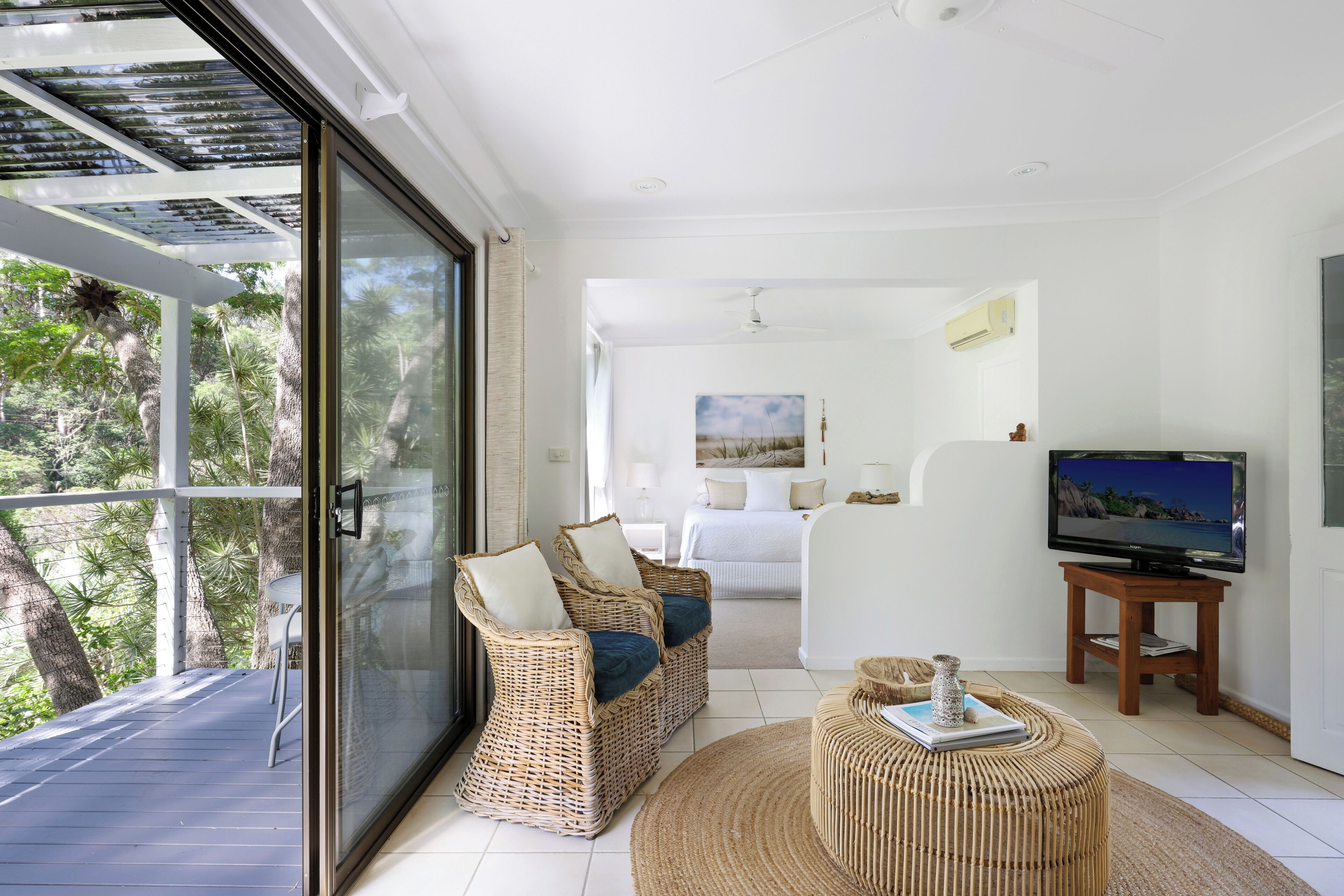 A modern living room with a large glass door leading to a balcony, wicker furniture, a television, and a bed in the background.