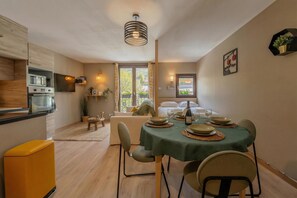 Une cuisine compacte dotée d’appareils électroménagers modernes, un coin repas avec une table dressée pour deux personnes, et un coin salon confortable avec un canapé et une petite table d’appoint.