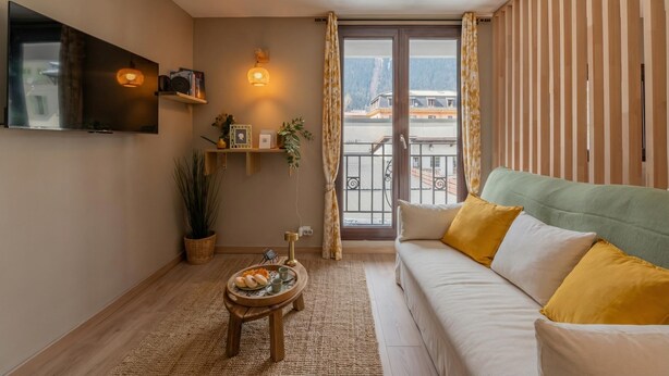 A living room with a sofa, a flat-screen TV, a small table with a tray of food, a potted plant, and a view of a building through a window.