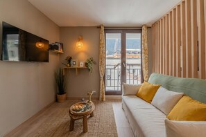 Un salon avec un canapé, un téléviseur à écran plat, une petite table sur laquelle se trouve un plateau de nourriture, une plante en pot et une vue sur un immeuble par la fenêtre.
