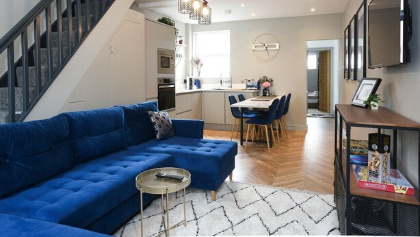 Una sala de estar moderna con un sofá seccional azul, un comedor con una mesa y sillas blancas, una cocina al fondo y una escalera que conduce a otro nivel.