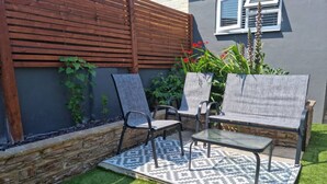 Un espacio al aire libre con dos sillas y una mesa, rodeado por un tapete estampado, frente a una pared con cerca de madera y plantas.