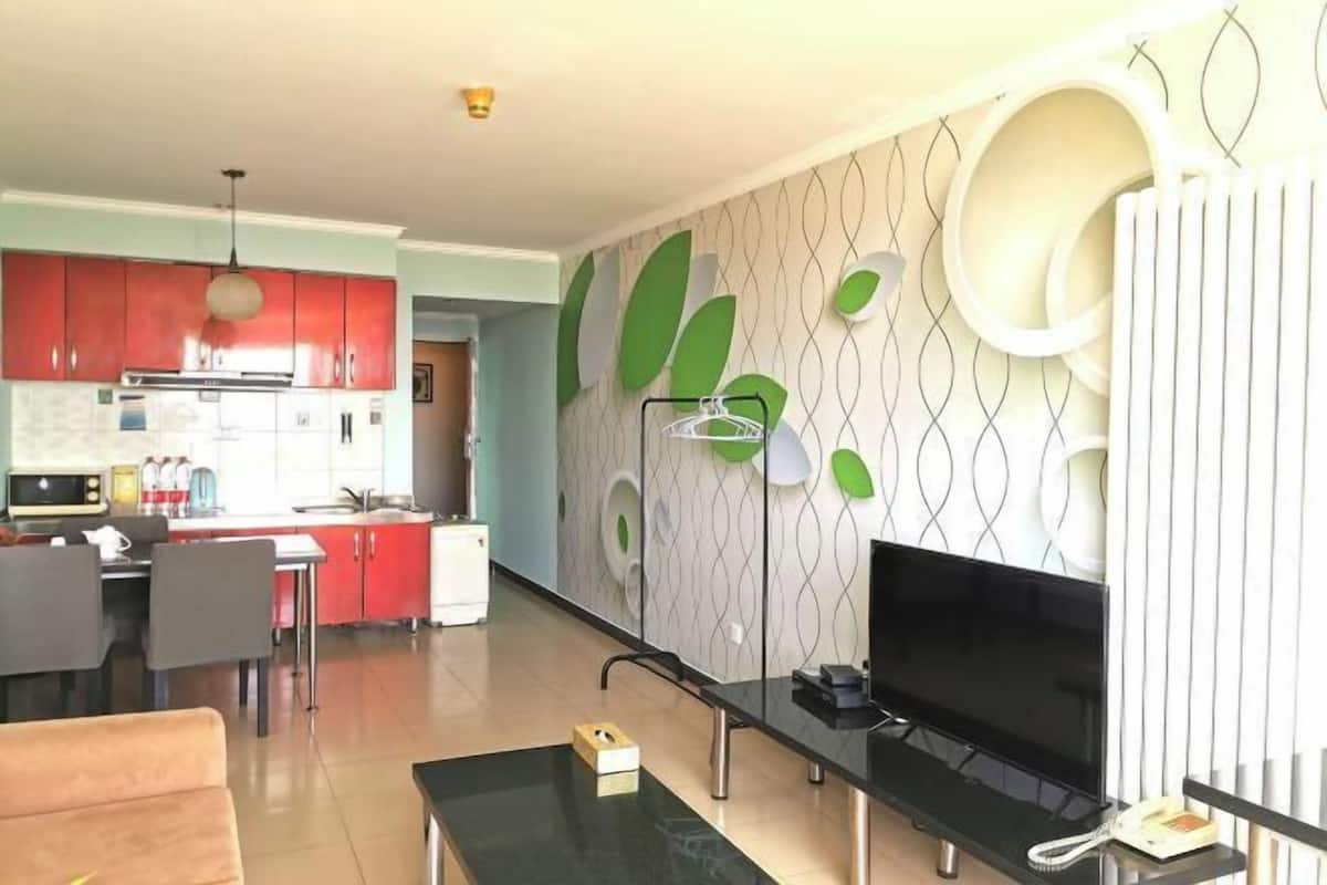 Een moderne keuken met rode kastjes, een eethoek met een tafel en stoelen, een woonkamer met een televisie, en een decoratieve wand met bladmotieven.