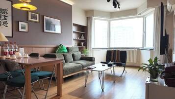 A modern living room with a sofa, a dining table with chairs, a coffee table, and a large window providing natural light.