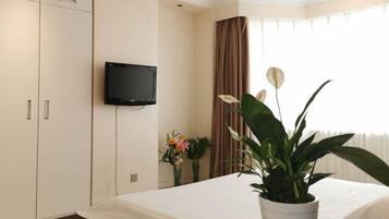 A hotel room with a bed, a TV mounted on the wall, a potted plant, and a window with curtains.